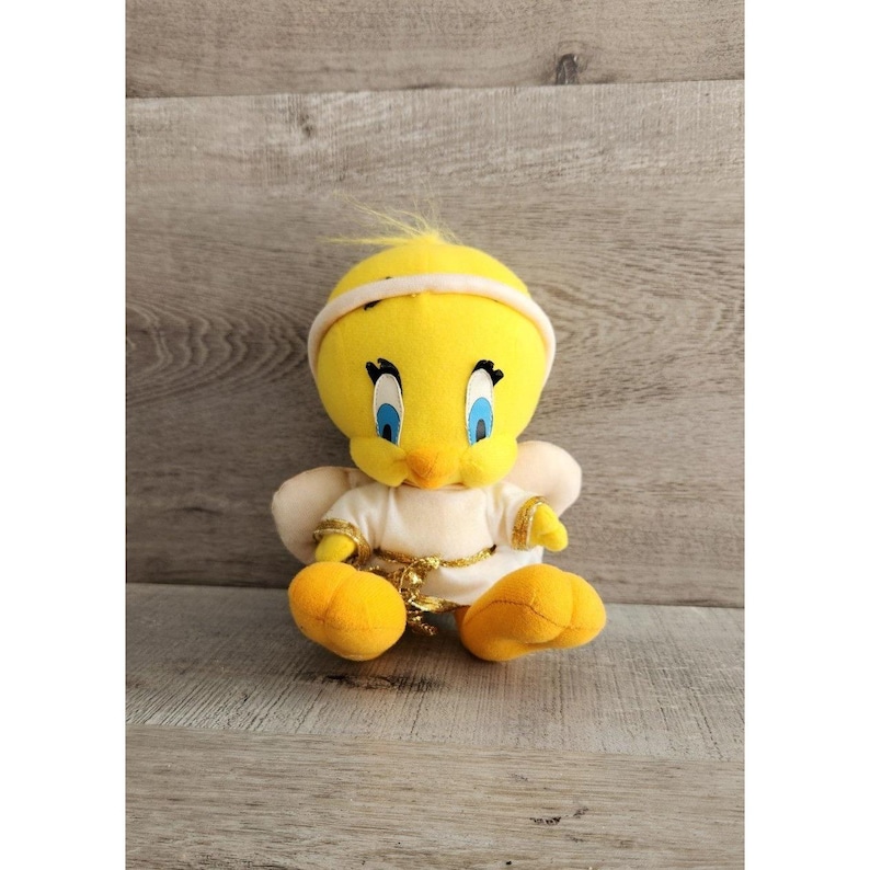 May include: A yellow plush Tweety Bird toy wearing a white angel costume with gold trim. The toy has blue eyes and a yellow beak.