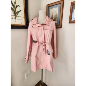 May include: A pink hooded jacket with a drawstring waist and embroidered details. The jacket has a zipper closure and two pockets.
