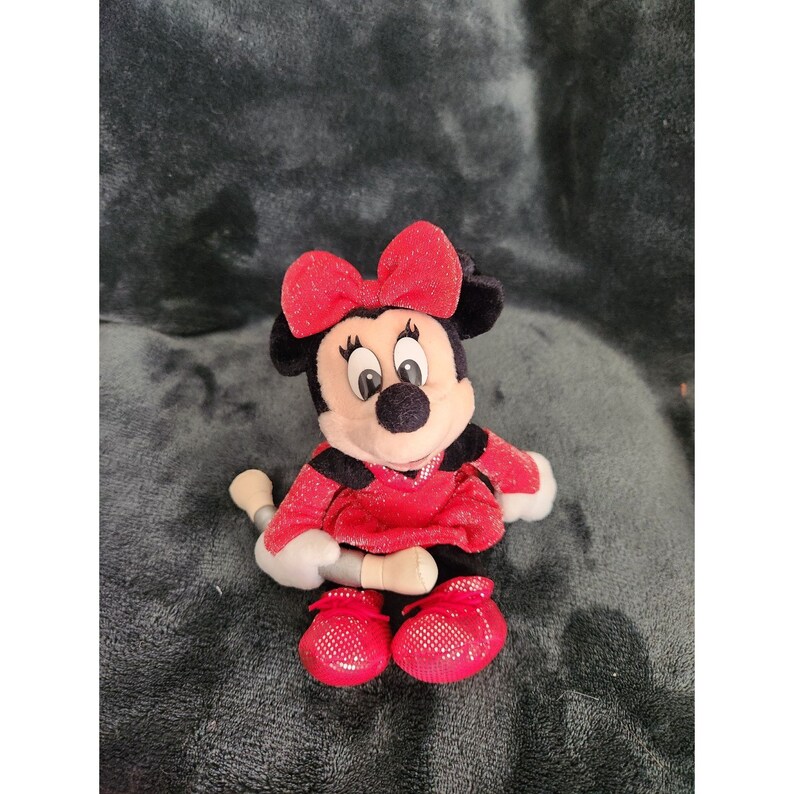 May include: A red plush Minnie Mouse toy with a sparkly red dress and a red bow. The toy is sitting on a gray fuzzy surface.