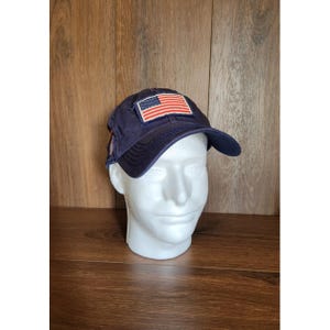 Pode incluir: Boné de beisebol azul marinho com um patch com a bandeira americana. O boné tem uma aba curva e é exibido em uma cabeça de manequim branca. O fundo é uma superfície de madeira.