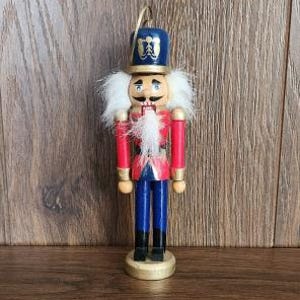 May include: A wooden nutcracker ornament featuring a blue hat with gold detailing, a red jacket, blue trousers, and black boots. The figure has white synthetic hair and a white beard, with a gold hanging loop attached to the top of the hat.