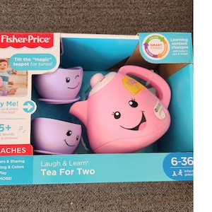 Op de afbeelding: Roze plastic theepot met een lachend gezicht en een geel theezakje. Twee paarse kopjes met lachende gezichten zijn inbegrepen. De doos zegt "Laugh & Learn Tea For Two" en "35+ liedjes, geluiden, melodieën en zinnen!"