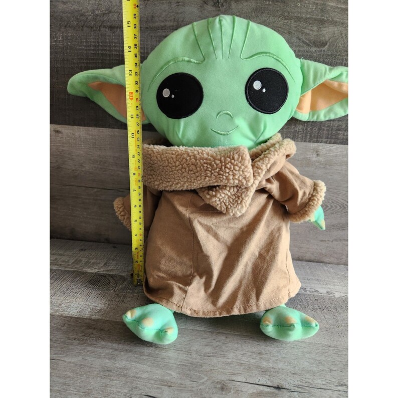 May include: Green plush toy of a character from the Star Wars franchise, wearing a brown robe. The toy is approximately 15 inches tall.
