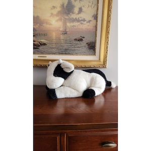 May include: A black and white plush cow laying on its side with its head resting on its front paws. The cow is on a dark brown wooden surface.