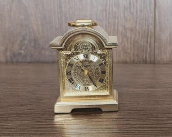 May include: A gold-toned mantel clock with an arched top and a ring handle. The clock face features black Roman numerals, gold hands, and the text Elgin and Quartz. The square frame has an intricate embossed floral pattern on the front.