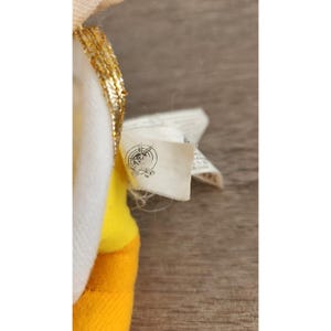 May include: A yellow and white plush toy with a gold trim and a white tag that says "Made in China" on it.