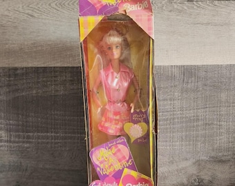 May include: A Barbie doll in a pink shirt and plaid skirt inside a tall cardboard box. The packaging features the text Make A Valentine, Barbie, and Cool stencils inside. The box is decorated with pink and yellow heart and plaid patterns.