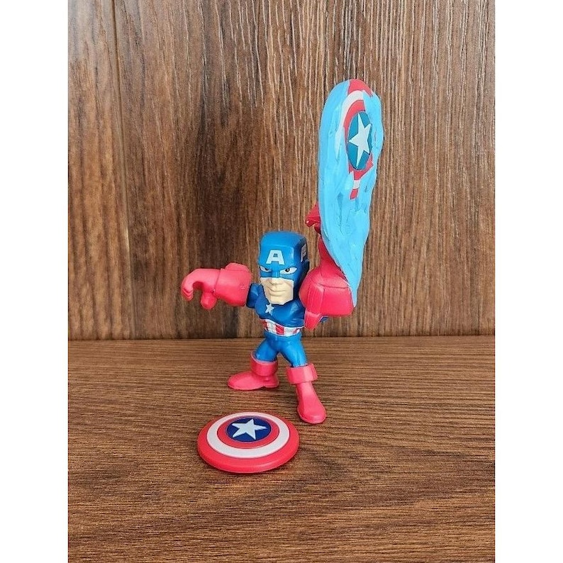 May include: A Captain America action figure in a dynamic pose, featuring a blue and red uniform, a shield, and a blue and red shield-shaped accessory. The figure is on a wooden surface.