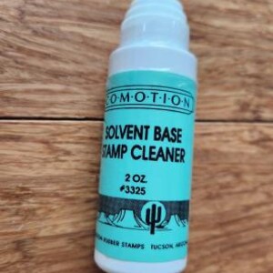 May include: A white plastic bottle of Co-Motion solvent base stamp cleaner with a light blue label. The label features the text "SOLVENT BASE STAMP CLEANER", "2 OZ.", "#3325", and a graphic of a cactus in a desert landscape.