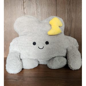 May include: A grey cloud-shaped plush pillow with a smiling face and a yellow accent. The pillow has a soft, fuzzy texture and a cute design, suitable for a child's room or as a decorative item.