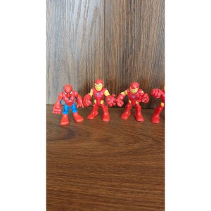 May include: A collection of colorful toy superhero figures, including Spider-Man and Iron Man, are arranged on a wooden surface. The figures are primarily red, blue, and yellow, with exaggerated features and a cartoonish design.