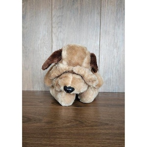 May include: A plush dog toy in shades of tan and brown. The toy has floppy ears, a black nose, and a soft, cuddly appearance. The dog is sitting on a wooden surface.