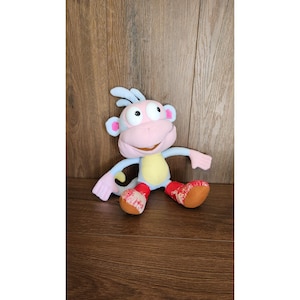 May include: A plush toy of a cartoon monkey with a pink face, blue arms and ears, and a yellow belly. The monkey wears red and brown shoes. The toy is sitting on a wooden surface.