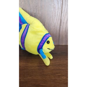 May include: A yellow plush fish toy with purple and blue stripes and a blue fin. The fish has black eyes and a smiling mouth.