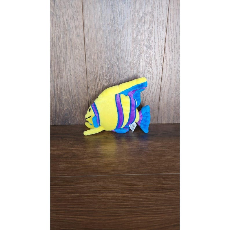 May include: A yellow plush fish toy with purple and blue stripes. The fish has a smiling face and a blue tail fin.