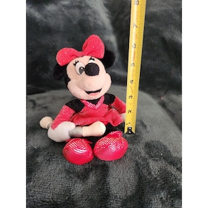 May include: A red and black plush Minnie Mouse toy holding a microphone. She is wearing a red sequined dress and a red bow. The toy is approximately 6 inches tall.