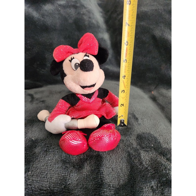May include: A red and black plush Minnie Mouse toy holding a microphone. She is wearing a red sequined dress and a red bow. The toy is approximately 6 inches tall.