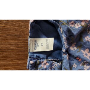 Puede incluir: Un vestido OshKosh B'Gosh de estampado floral azul y rosa con una etiqueta blanca que dice "OshKosh B'Gosh" y "2T" y "Made in Sri Lanka Fabrique au Sri Lanka Hecho en Sri Lanka".