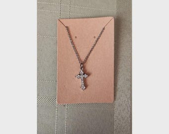 May include: A silver-toned metal necklace featuring a cross pendant encrusted with small clear stones. The necklace is displayed on a rectangular light brown cardboard backing card with two small holes at the top for the chain.