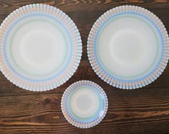 May include: Two large round plates and one smaller round plate made of white milk glass. Each plate features a scalloped edge and a decorative border pattern consisting of alternating thin stripes in soft shades of blue, green, and peach.