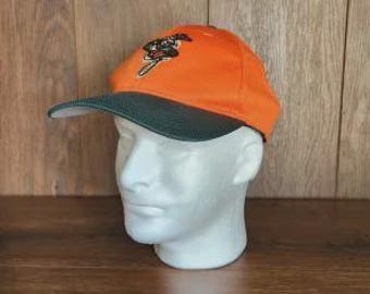 May include: A bright orange baseball cap with a dark green brim. The front of the cap features an embroidered graphic of a stylized figure in black, white, and red colors. The cap is displayed on a white mannequin head to show its shape.