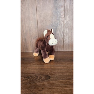 May include: A brown plush horse toy with a white face and brown hooves. The horse is standing on a wooden surface.