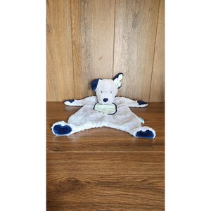 May include: A soft, dog-shaped baby comforter in light grey with navy blue details on the ears, paws, and edges. It features a light green patch on the chest and is placed on a wooden surface.