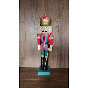 May include: A wooden nutcracker figurine, painted in red, teal, and gold, stands on a teal base. The figurine wears a gold crown and holds a golden sword. The nutcracker has a white mustache and a stern expression. The figurine is approximately 12 inches tall.
