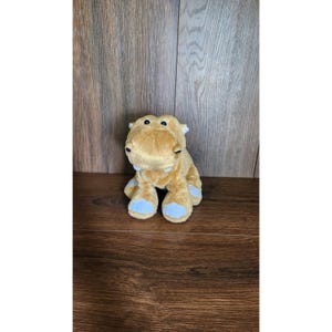 May include: A plush hippo toy in a sitting position. The hippo is primarily tan with light blue accents on its feet and ears. It has small black eyes and a soft, cuddly appearance, perfect for children.