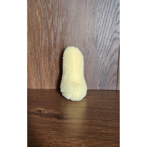 以下が含まれることがあります： 小さくふわふわとした黄色のぬいぐるみで、おもちゃや装飾品かもしれません。丸みを帯びたベースと細長い上部があり、柔らかく質感のある外観です。木製の背景に配置されています。