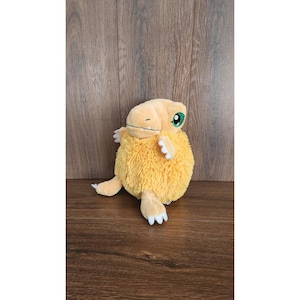 Puede incluir: Un peluche amarillo de una criatura con cuerpo redondo, brazos y piernas pequeños. La criatura tiene una cabeza amarilla clara con un ojo cerrado y un ojo verde. El peluche está hecho de material suave y difuso.