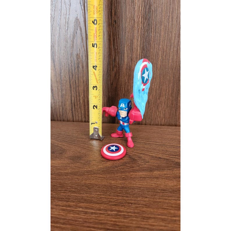 May include: A Captain America action figure, approximately 3 inches tall, with a blue and red costume, holding a shield. A separate red and white shield is also present. The figure is on a wooden surface.