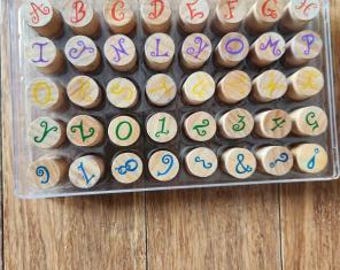 Könnte beinhalten: Ein durchsichtiger Kunststoffbehälter mit vierzig zylindrischen Holzstempeln. Jeder Stempel zeigt einen farbigen Buchstaben, eine Zahl oder ein Symbol auf der Oberseite in einer geschwungenen Schriftart, angeordnet in fünf Reihen.