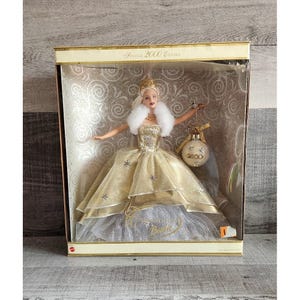 Może przedstawiać: Lalka Barbie Special 2000 Edition w oryginalnym pudełku. Lalka ma na sobie złotą suknię z białym futrzanym szalem i koroną. Pudełko ma złoty i biały wzór z napisem "Special 2000 Edition".