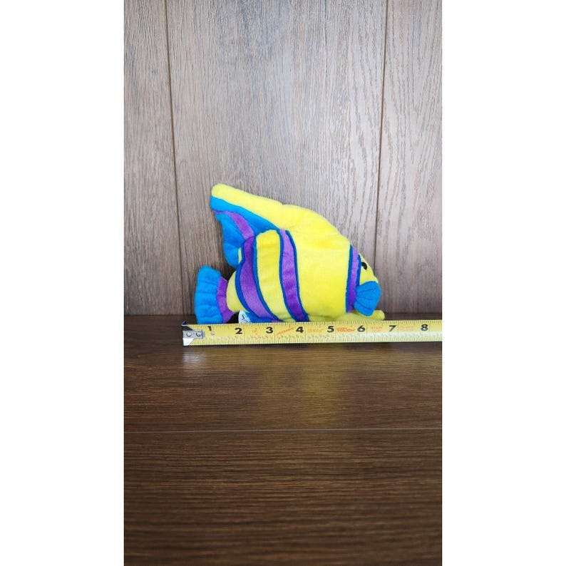 May include: A yellow and blue plush fish toy with purple stripes. The fish is about 5 inches long.