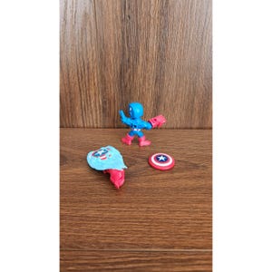 May include: A Captain America action figure with a blue helmet and suit, red gloves and boots, and a shield. The figure is posed with its shield and a separate shield piece and a round shield with a star.