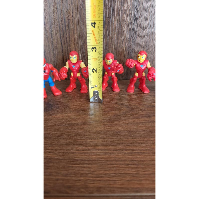 May include: A collection of small, red and yellow Iron Man action figures, each approximately 2 inches tall, are arranged on a wooden surface. A Spider-Man figure is also present. A yellow measuring tape is placed next to the figures.