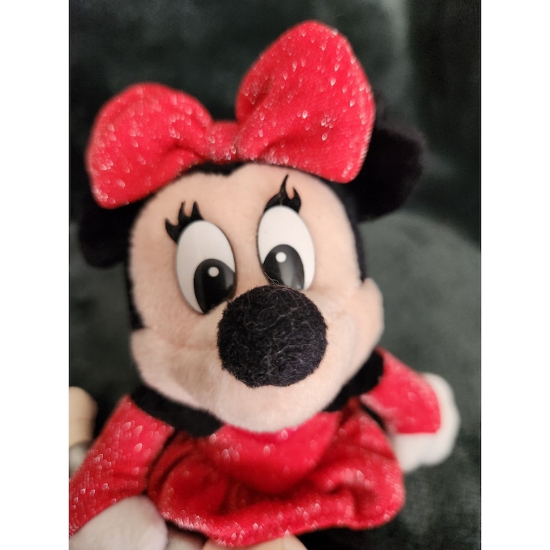 May include: A red plush toy with a black nose and large eyes. The toy is wearing a red bow and a red dress with white polka dots.