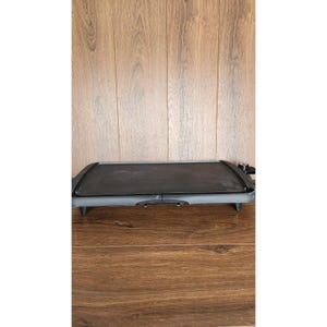 Könnte beinhalten: Eine rechteckige elektrische Grillplatte mit antihaftbeschichteter Kochfläche. Die Grillplatte ist schwarz mit einem grauen Rahmen und steht auf kurzen Beinen. Ein Temperaturregler ist an der Seite sichtbar. Die Grillplatte ist für das Kochen in Innenräumen konzipiert.