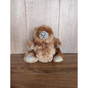 May include: A brown plush teddy bear with a grey nose and black eyes. The bear is sitting on a wooden surface.