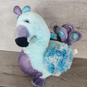 Puede incluir: Un pájaro de peluche con pelaje azul claro, vientre de malla morada y pico negro. Tiene detalles en morado y turquesa en la cabeza y la cola, con texturas de tela en la cresta y las plumas de la cola. El juguete tiene un cuerpo suave.