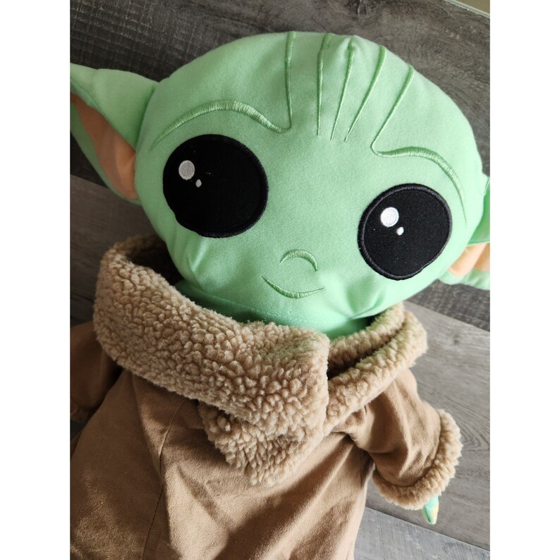 May include: Green plush toy with large eyes and a brown fur-lined coat. The toy is a popular character from the Star Wars franchise.