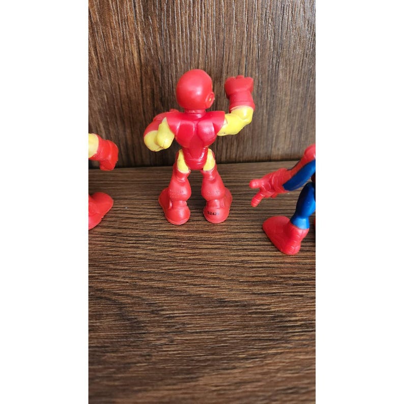 May include: A red and yellow Iron Man action figure with raised fist, part of a set of superhero toys. The figure is made of plastic and has a cartoonish design. Other figures are partially visible.