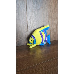 May include: Yellow and purple plush fish toy with blue fins. The fish has a smiling face and is lying on a wooden surface.