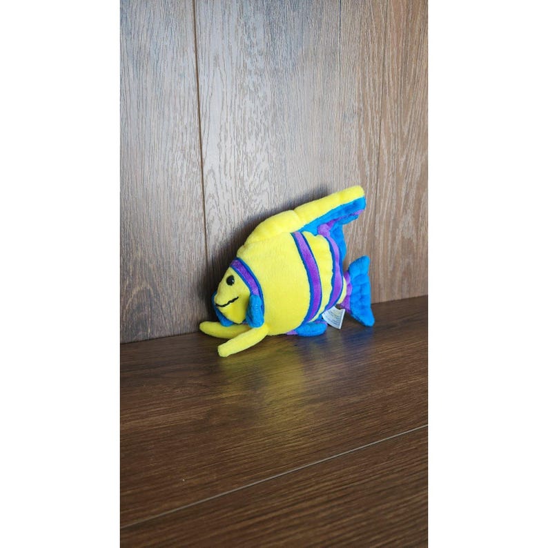 May include: Yellow and purple plush fish toy with blue fins. The fish has a smiling face and is lying on a wooden surface.