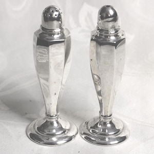 May include: A pair of silver salt and pepper shakers. Each shaker has a faceted, elongated body, a circular base, and a rounded top. One shaker has holes on the top, and the other has a solid top. They are on a white surface.