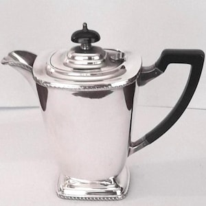 May include: A silver-coloured coffee pot with a polished metallic finish, featuring a black handle and a matching black finial on the lid. The vessel has a square base with a decorative rope-style border and a curved spout on the left side.