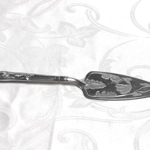 May include: Silver cake server with a decorative handle and an etched floral design on the blade. The server is made of shiny metal and is set against a white background with a floral pattern.