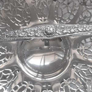 May include: A silver-coloured metal serving dish featuring an intricate openwork filigree pattern along the scalloped edges. A horizontal handle crosses the centre, decorated with a detailed embossed floral and leaf motif in the middle.