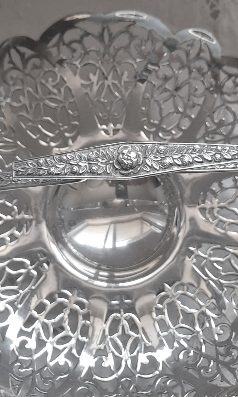 May include: A silver-coloured metal serving dish featuring an intricate openwork filigree pattern along the scalloped edges. A horizontal handle crosses the centre, decorated with a detailed embossed floral and leaf motif in the middle.